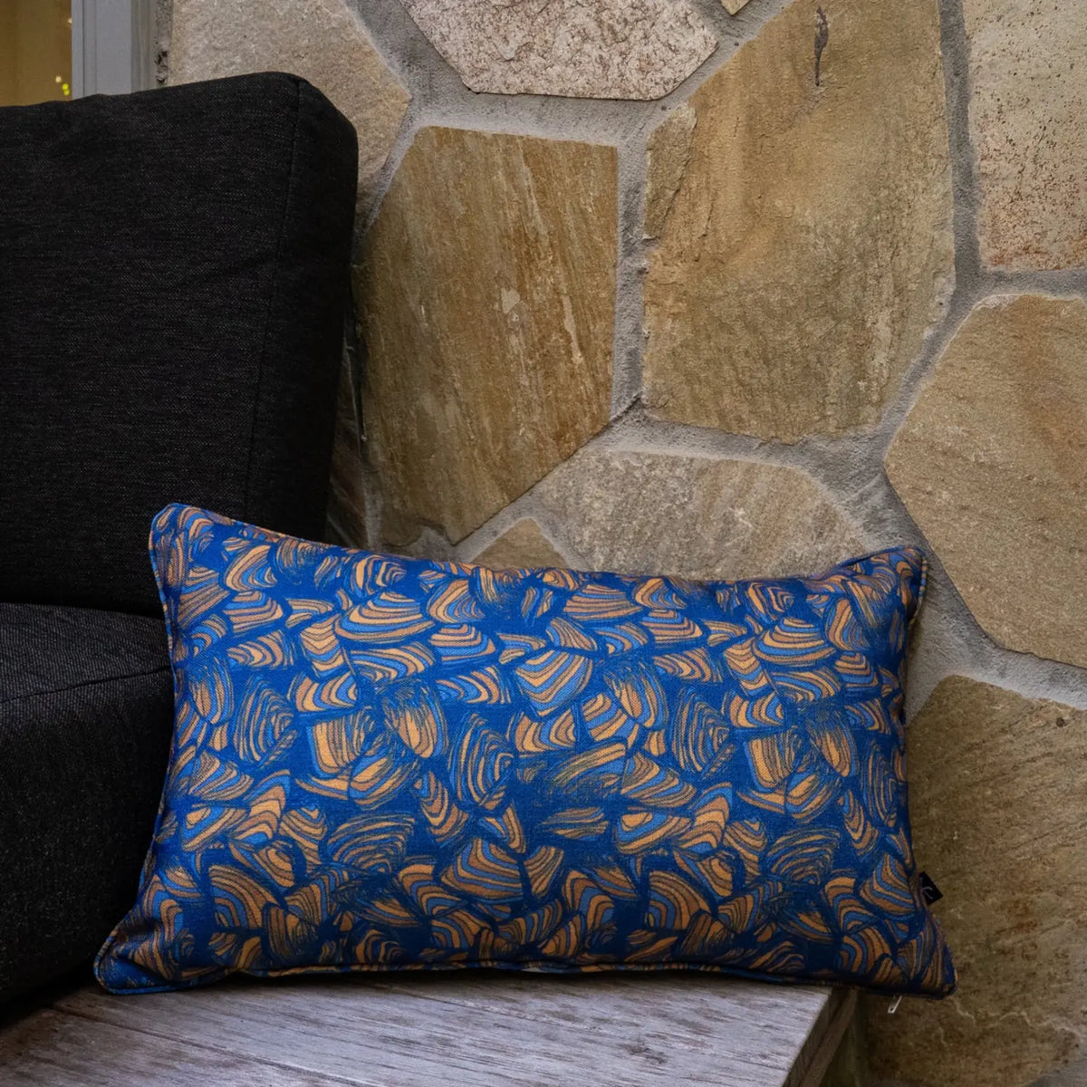 Lumbar Pipi Mariner cushion with blue and tan abstract print styled against a stone wall.