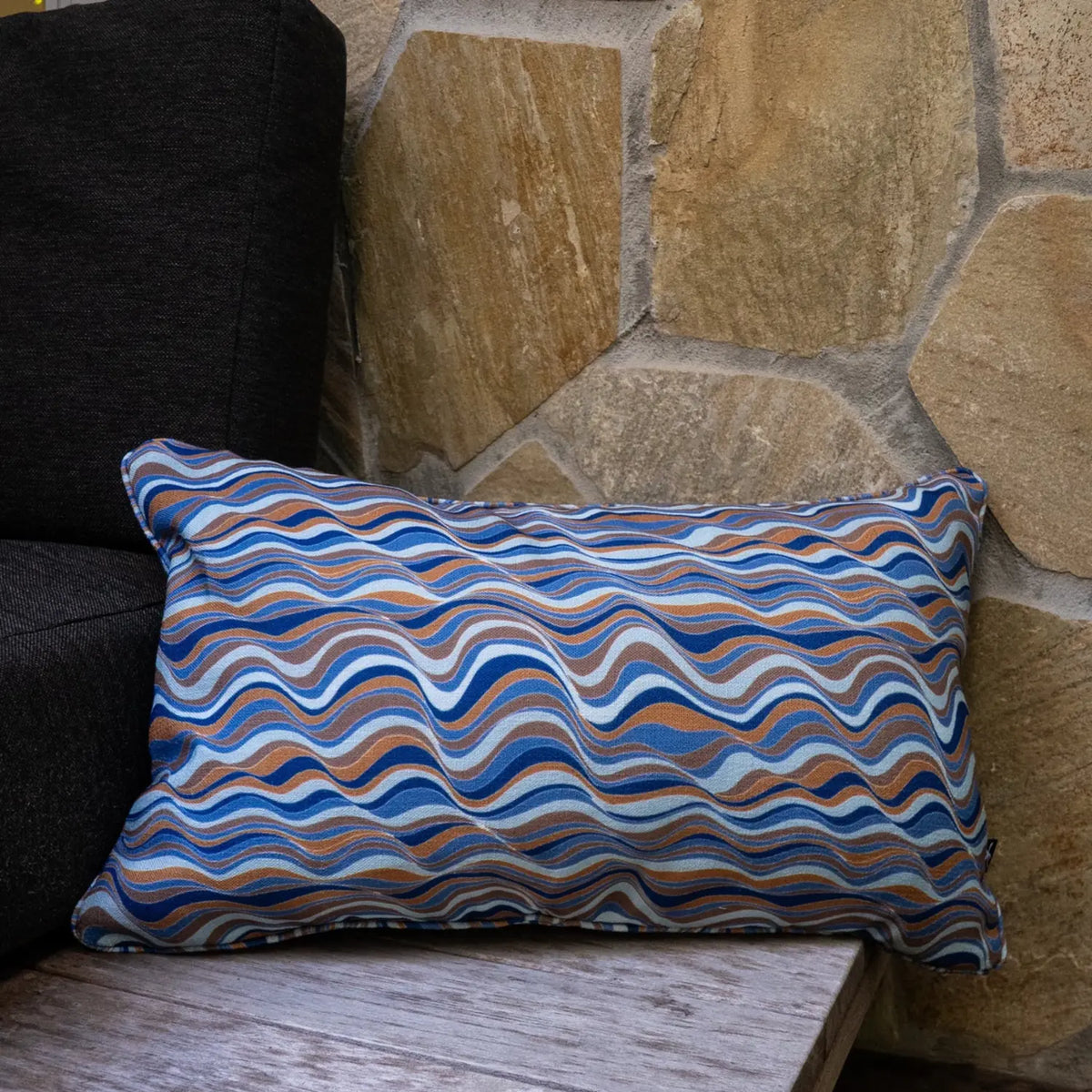 Lumbar straddie stripes outdoor cushion with flowing blue and tan wave design styled against a stone wall.