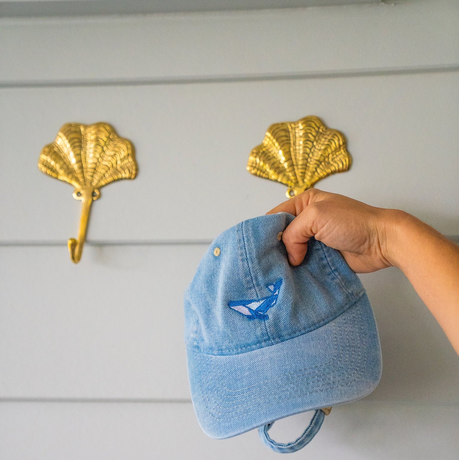 Hand holding a blue denim cap above gold pineapple hooks on a white wall.