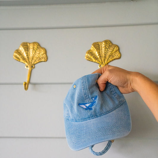 Hand holding a blue denim cap above gold pineapple hooks on a white wall.