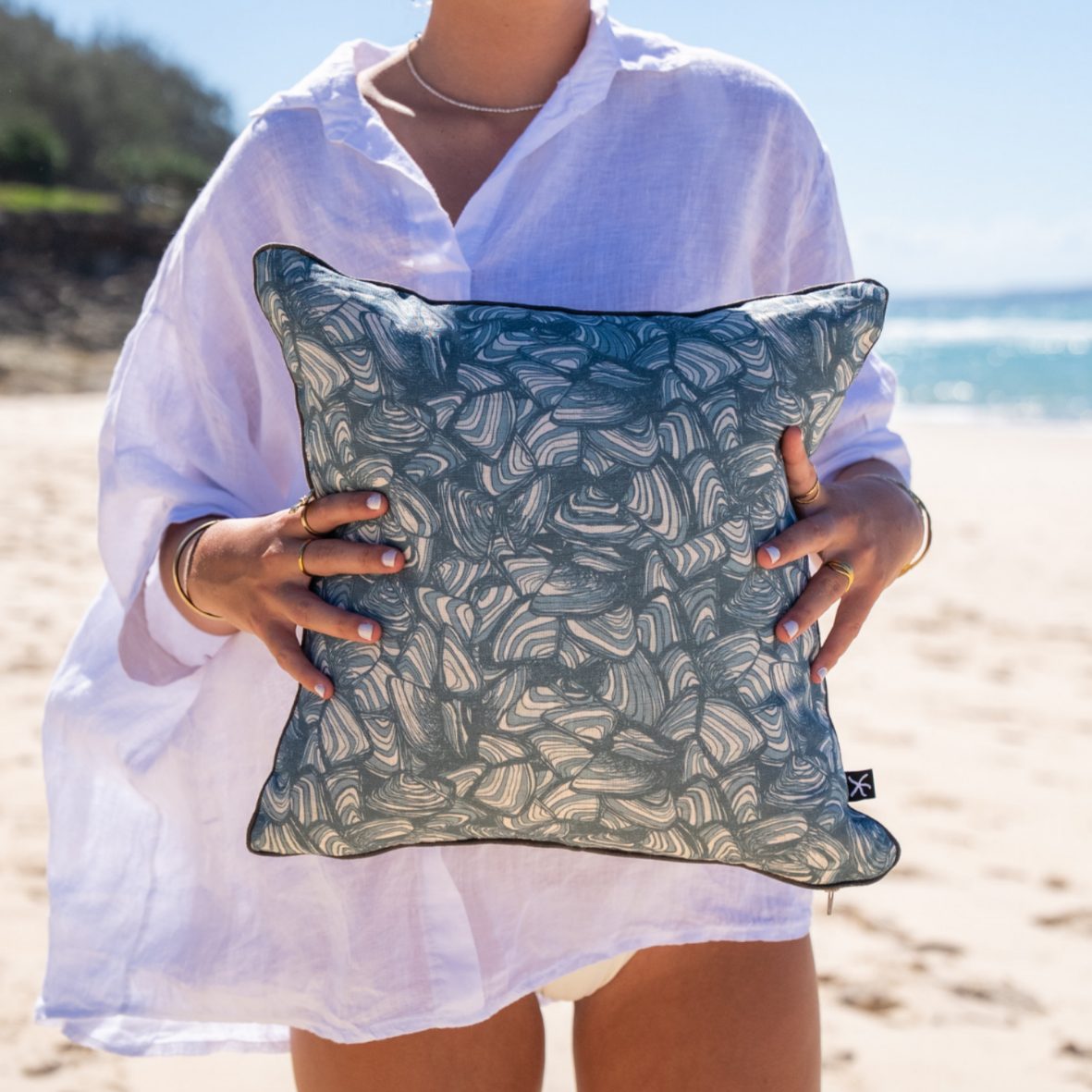 Woman holding a square linen cushion cover in the Frenchmans Pipi design on the beach.