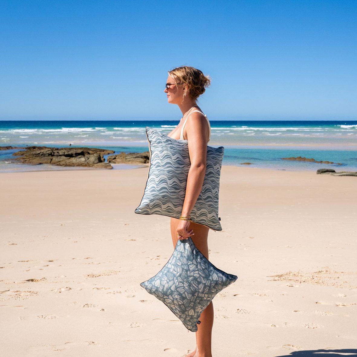 Side view of a woman walking along the beach carrying Frenchmans Pipi linen cushion covers.