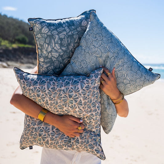 Woman holding three linen cushion covers on the beach, featuring the Frenchmans Shells design with coordinating Starfish Studio prints.