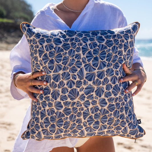 Mariner Shells linen cushion cover with blue shell print and ochre piping held on the beach