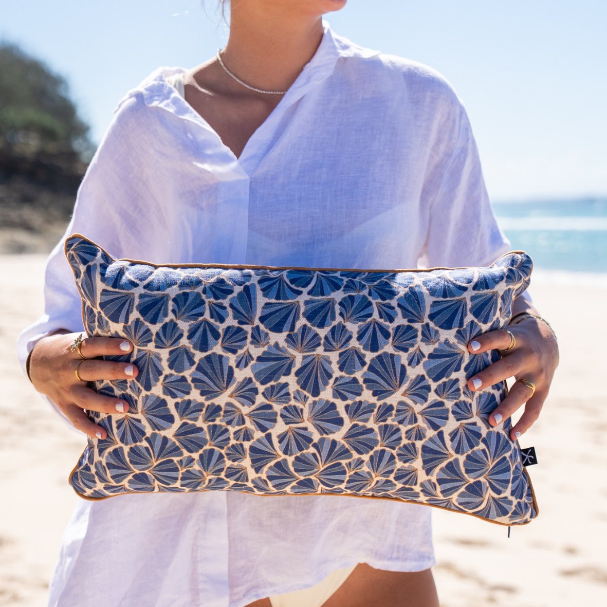 Mariner Shells rectangular linen cushion cover in blue and natural tones held on the beach