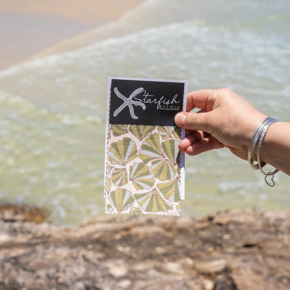 Shells fabric swatch by Starfish Studio showing the green shell-inspired print against a coastal water background.