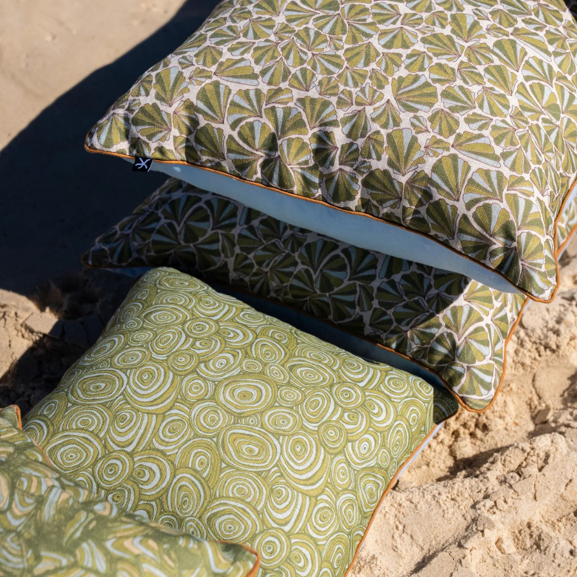 Stack of Shells and Growth Rings linen cushion covers on the beach, showing green coastal prints and orange piping.