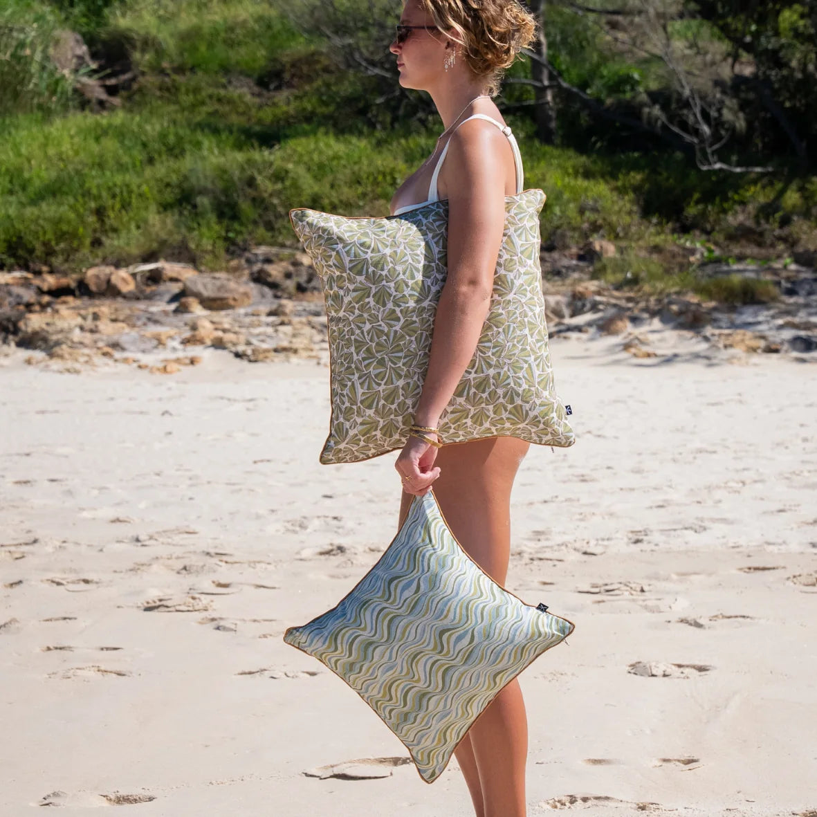 Woman carrying a green Shells linen cushion cover on the beach with a coordinating Straddie Stripe cushion.