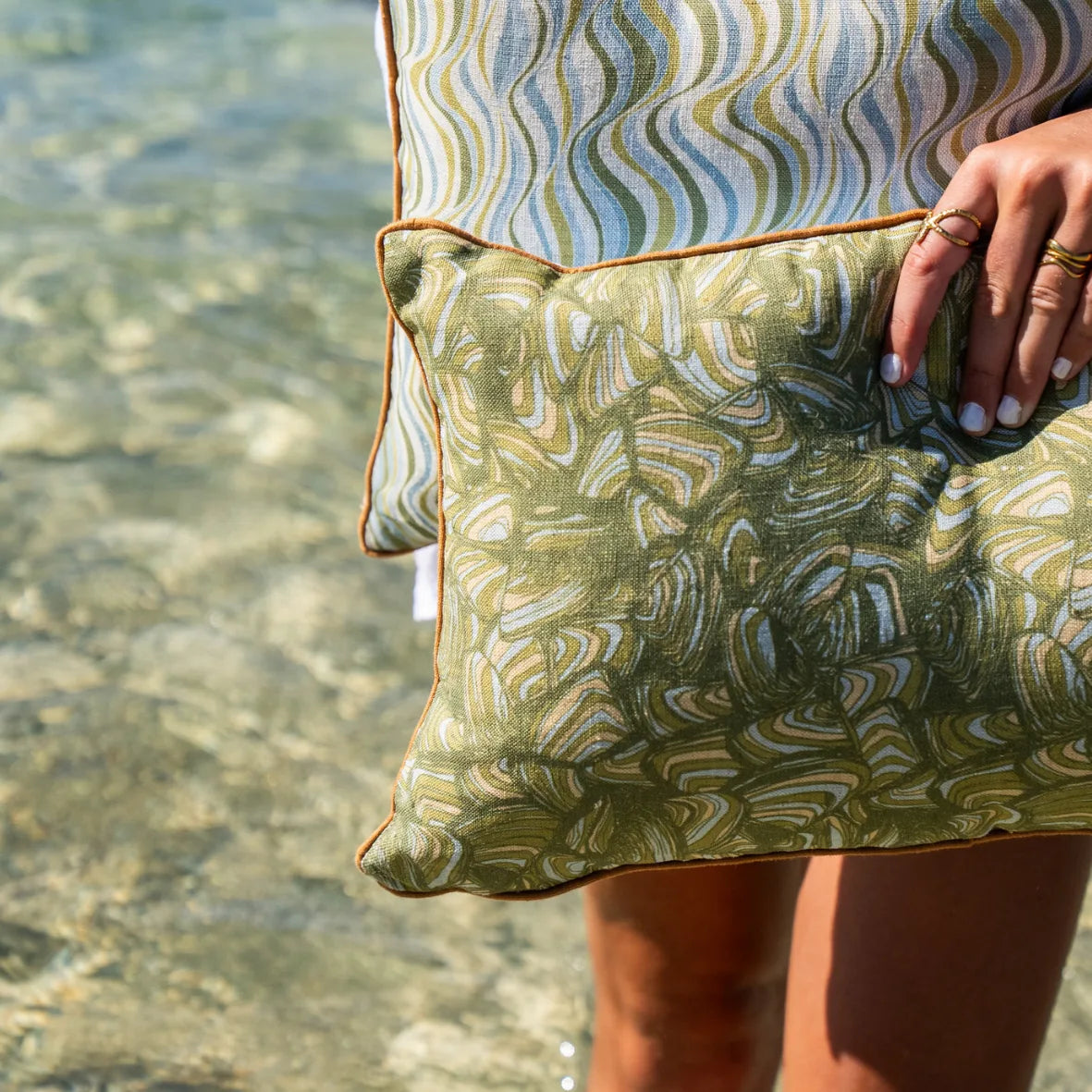 Close-up of olive green Pipi linen cushion cover showing the abstract print, orange piping and fabric texture beside clear coastal water.