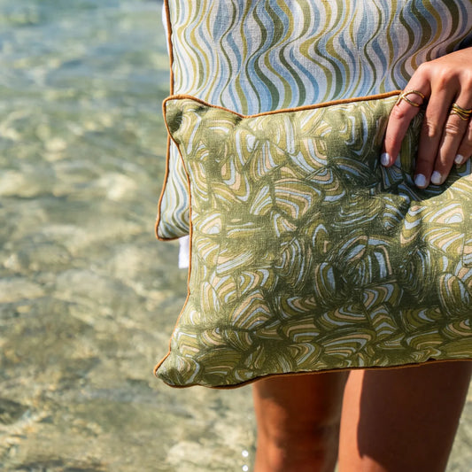 Close-up of olive green Pipi linen cushion cover showing the abstract print, orange piping and fabric texture beside clear coastal water.