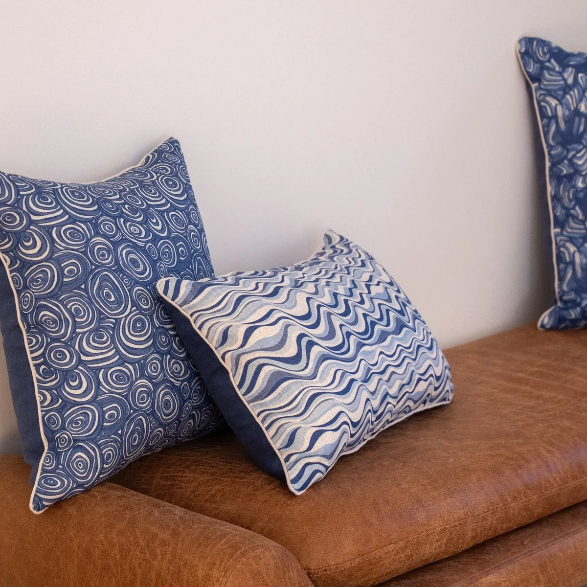 Pacific Pipi linen cushion cover styled with coordinating blue coastal cushions on a tan leather lounge