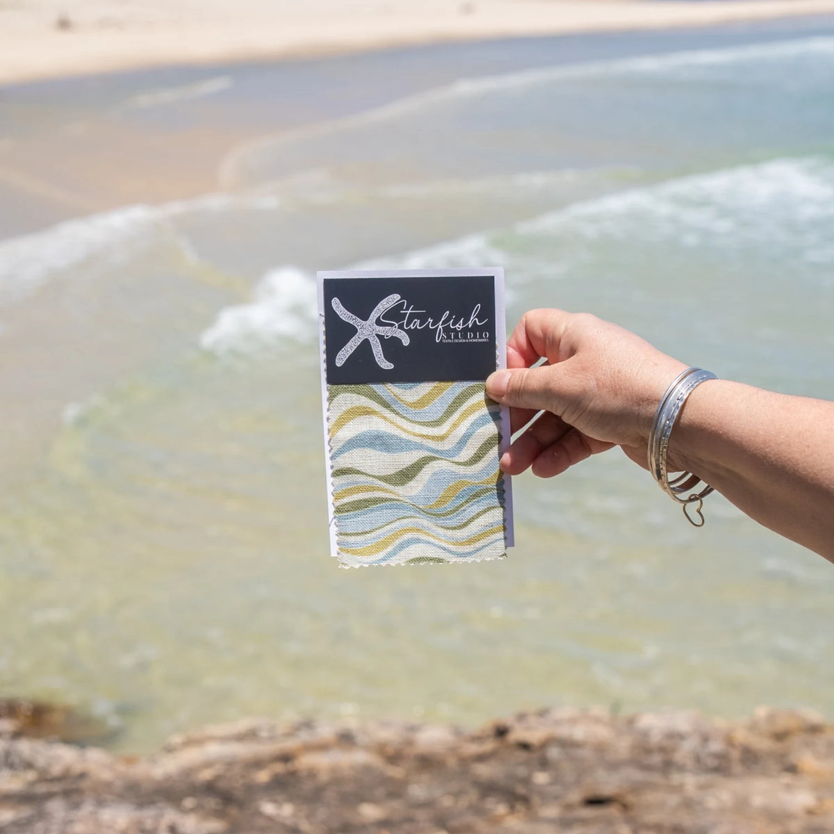 Straddie Stripe linen cushion cover fabric swatch held beside the ocean on North Stradbroke Island