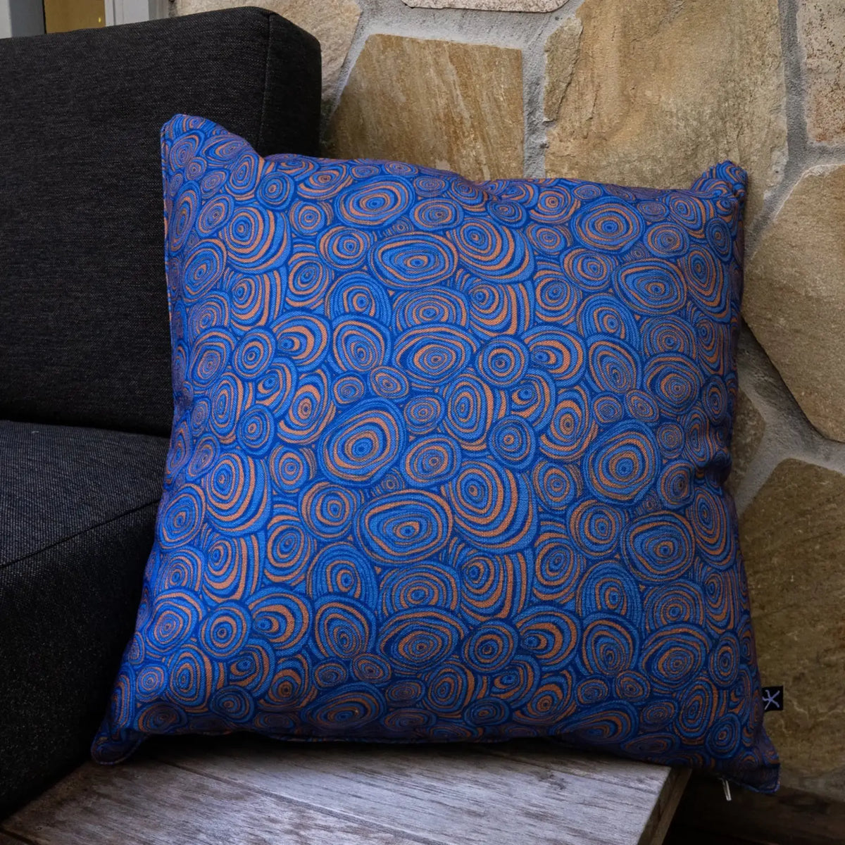 Square outdoor and indoor cushion in the Mariner colourway featuring the Growth Rings pattern, styled on a timber chair beside a stone wall.