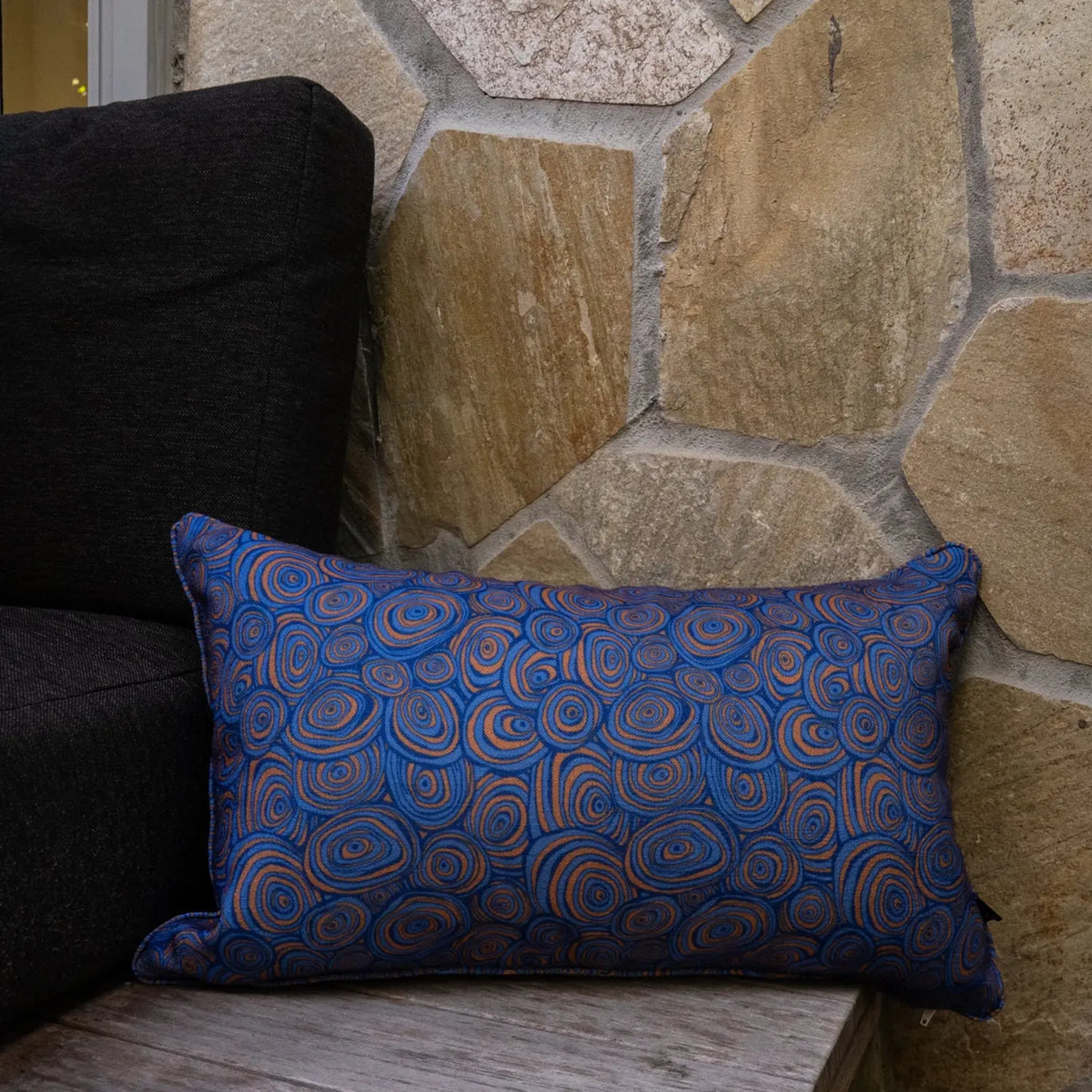 Lumbar outdoor and indoor cushion in the Mariner colourway featuring the Growth Rings pattern, styled on a timber chair beside a stone wall.