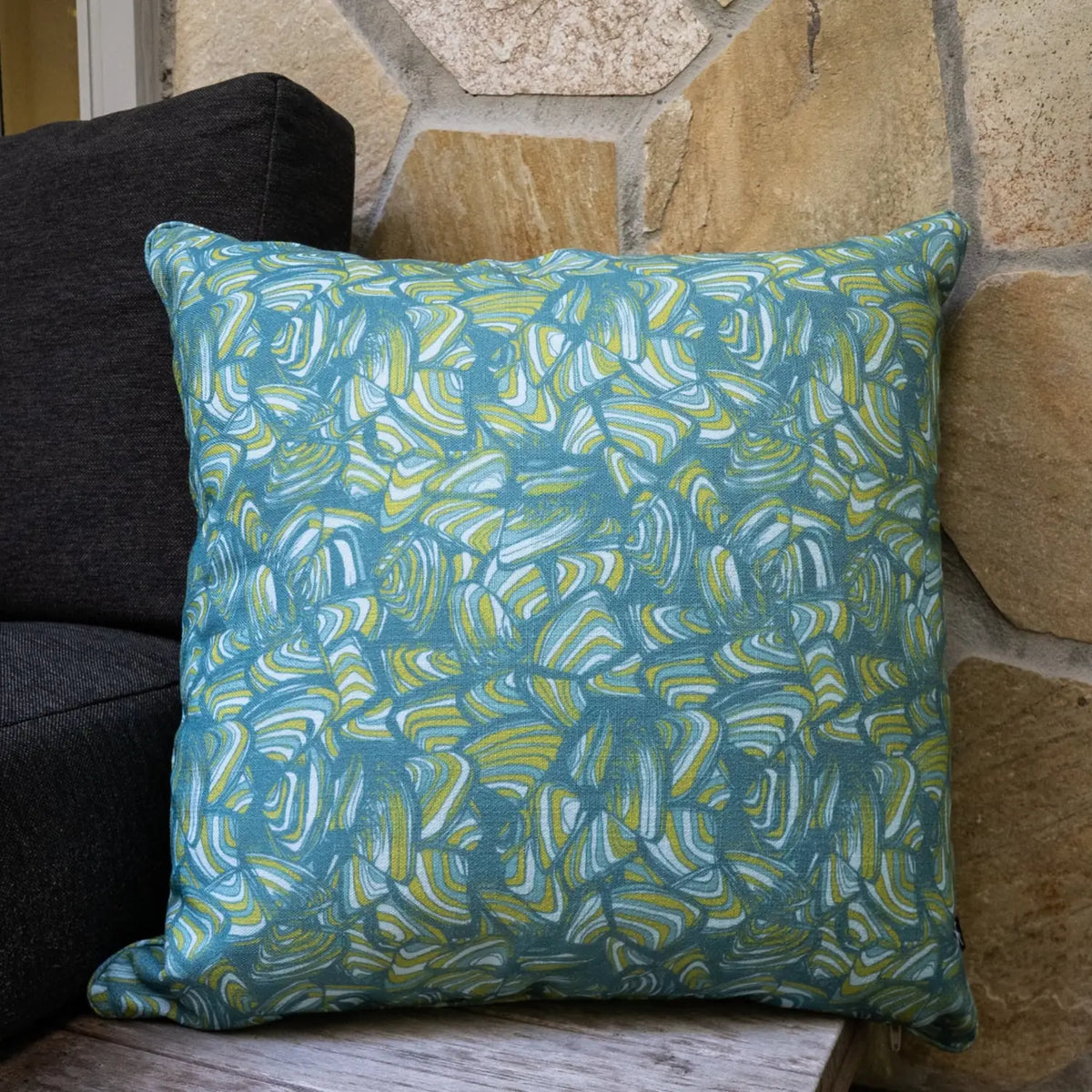 Square Mangrove Pipi cushion with blue, green and white abstract print styled against a stone wall.