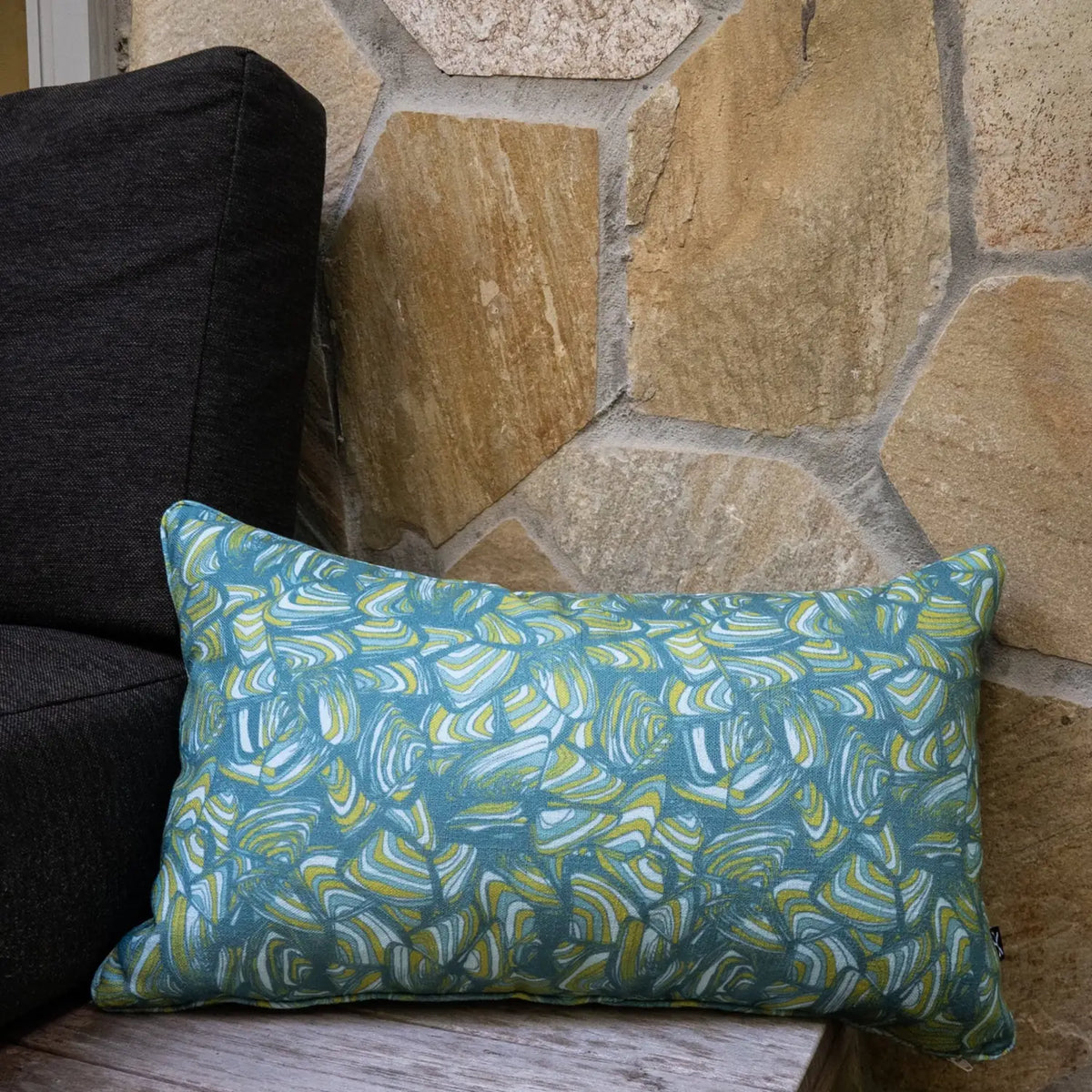 Lumbar Mangrove Pipi cushion with blue, green and white abstract print styled against a stone wall.