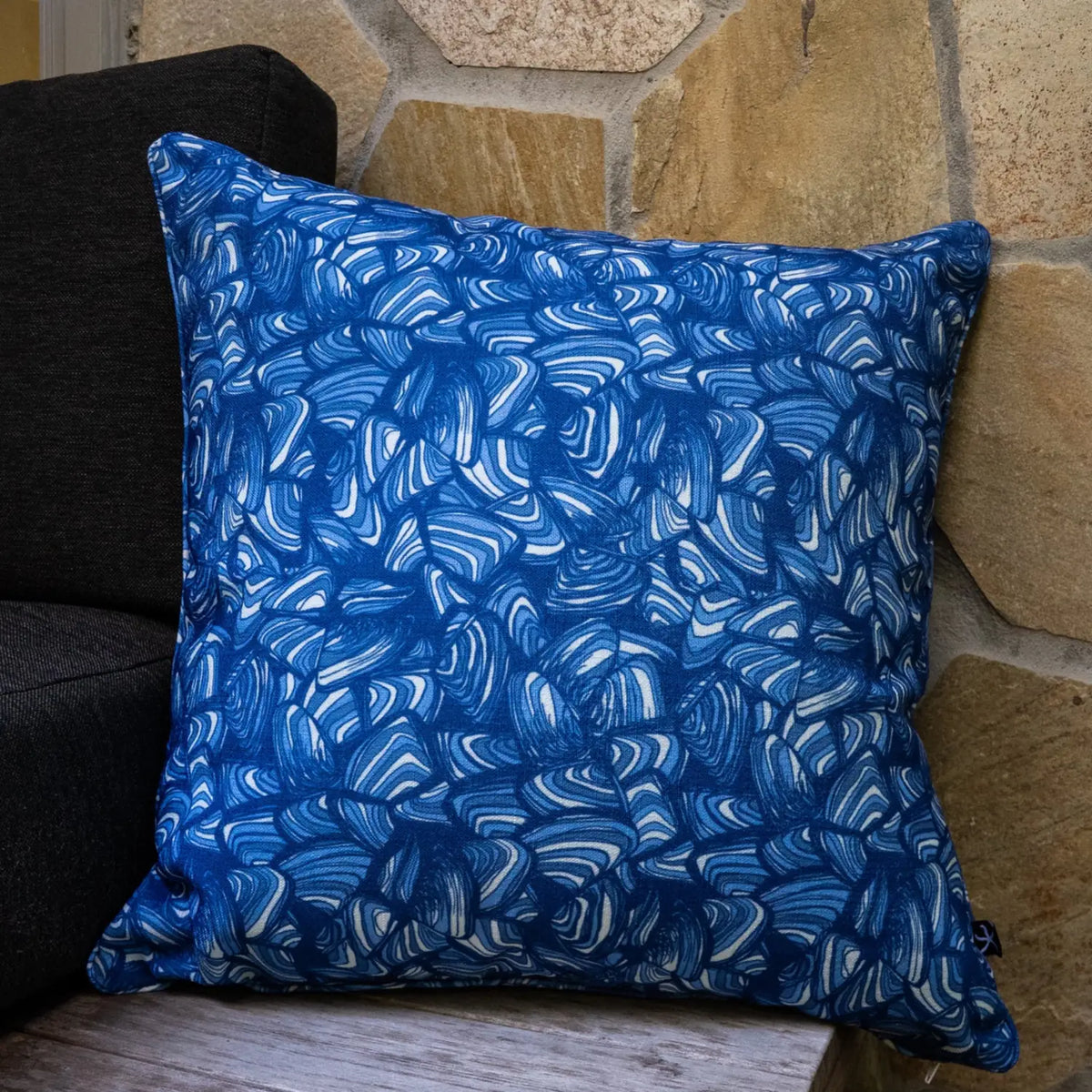 Pacific Pipi square cushion in blue and white abstract print styled against a stone wall.
