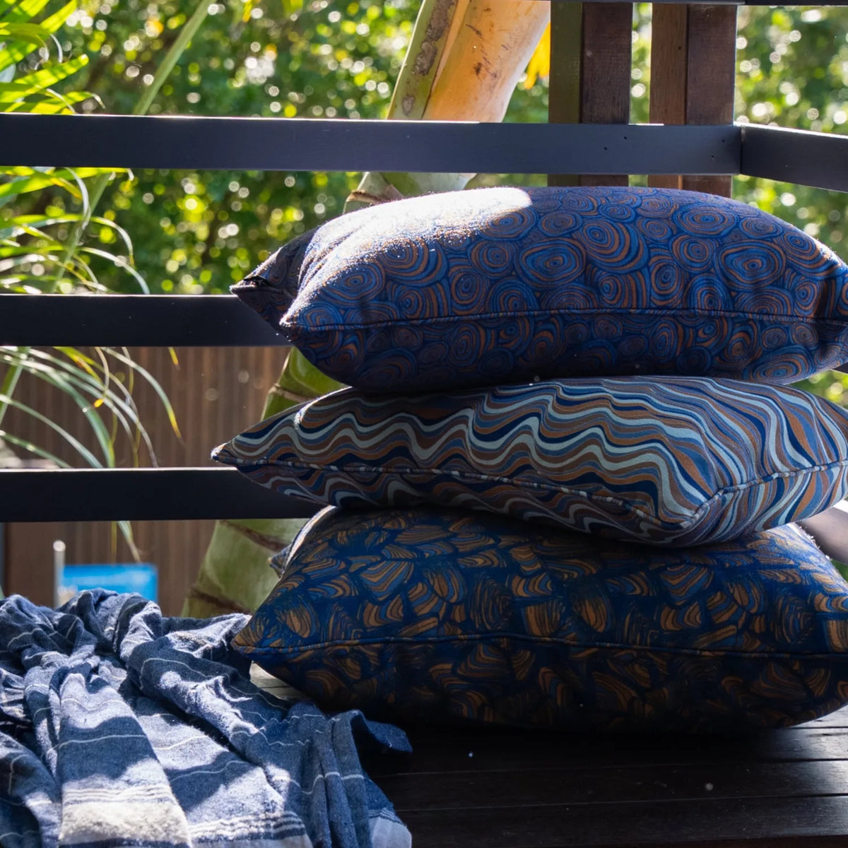 Stack of three patterned outdoor cushions styled on a timber deck with a blue striped throw.