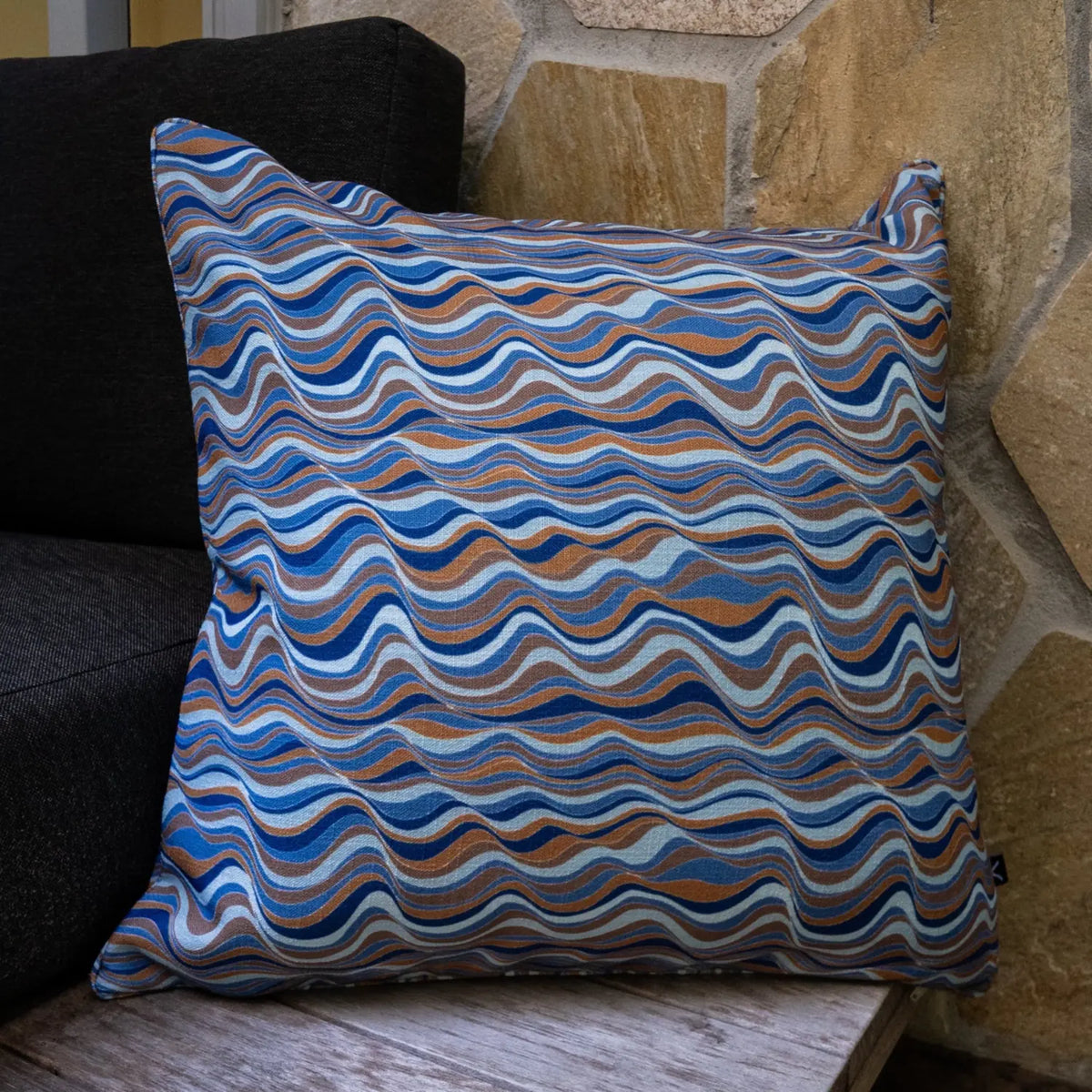 Square patterned outdoor cushion with flowing blue and tan wave design styled against a stone wall.